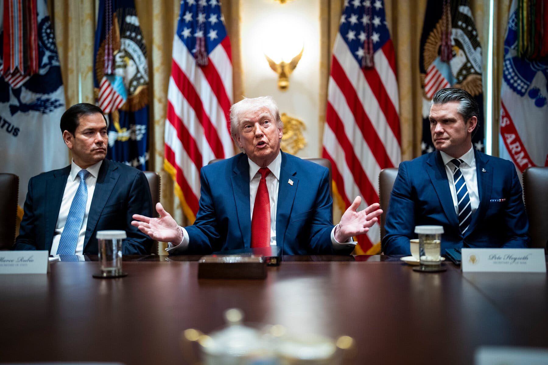 Marco Rubio, Donald Trump and Pete Hegseth sit behind a wooden table with American and military flags behind them.