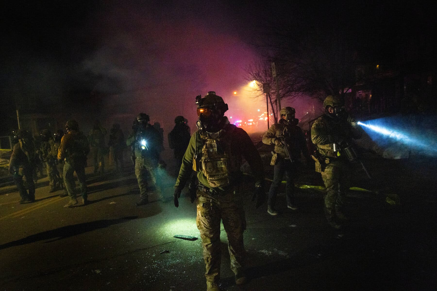 Armed individuals in tactical gear and gas masks walk through a dark, smoky street illuminated by flashlights.